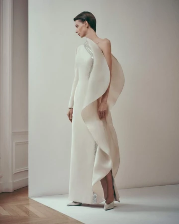 Elegant woman in a white gown poses in a studio. The gown has a one-shoulder design with draped fabric and embellishments.