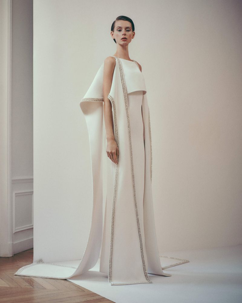 An adult female fashion model stands in a photo studio wearing an off-white gown with cape and column silhouette in a full shot.