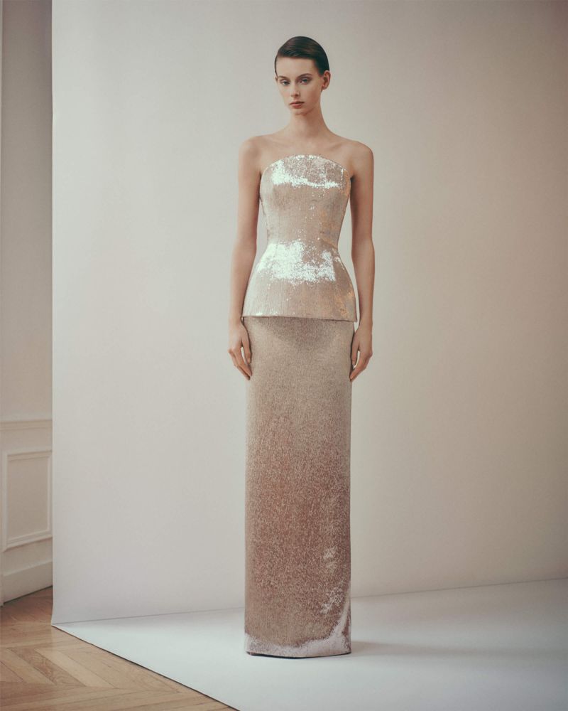An adult female fashion model poses in a sequined peplum gown in a photo studio with neutral walls and a hardwood floor, showcasing luxury style.
