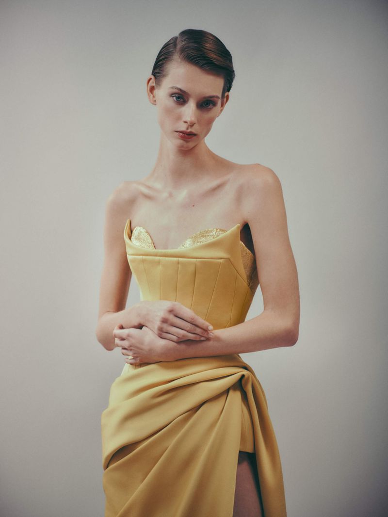 A slender woman poses in a yellow strapless dress with a draped skirt and a thigh-high slit against a plain background in a portrait.