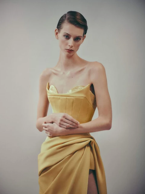 A slender woman poses in a yellow strapless dress with a draped skirt and a thigh-high slit against a plain background in a portrait.