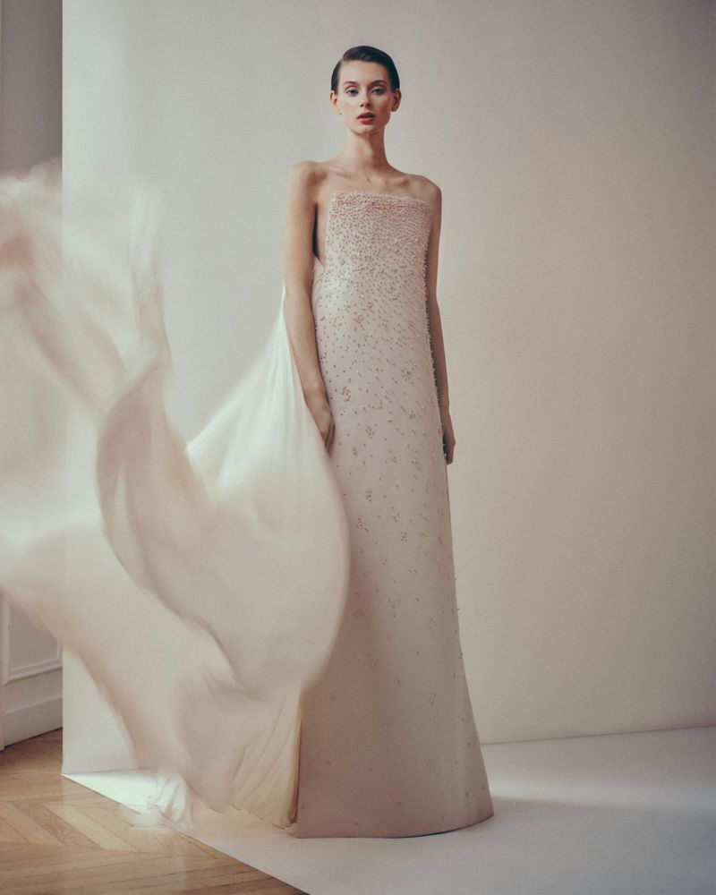 A woman wearing a light pink formal dress with beaded details poses in a studio with a large veil fluttering to the left.