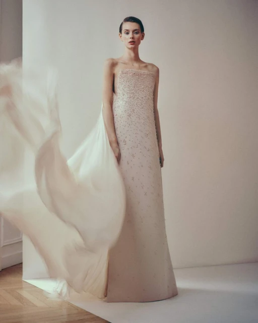 A woman wearing a light pink formal dress with beaded details poses in a studio with a large veil fluttering to the left.