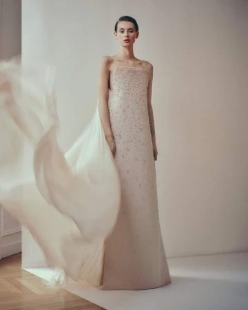A woman wearing a light pink formal dress with beaded details poses in a studio with a large veil fluttering to the left.