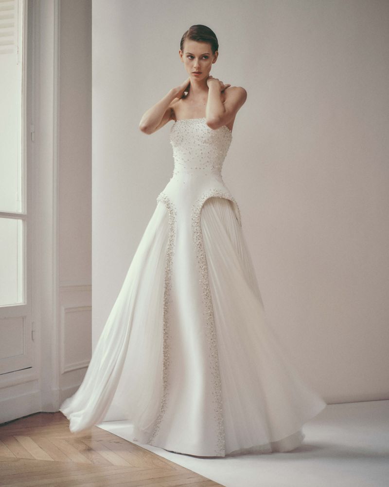 Elegant woman poses in a white strapless wedding gown with beadwork and tulle skirt in a minimalist studio setting with soft lighting.