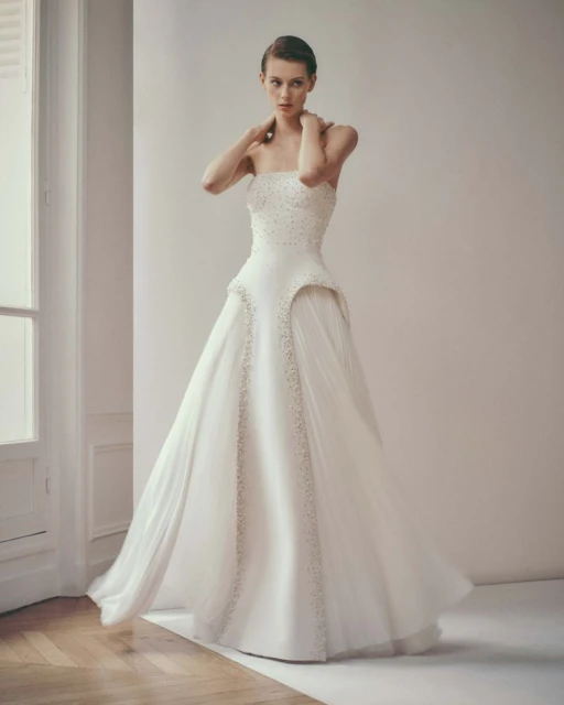 Elegant woman poses in a white strapless wedding gown with beadwork and tulle skirt in a minimalist studio setting with soft lighting.