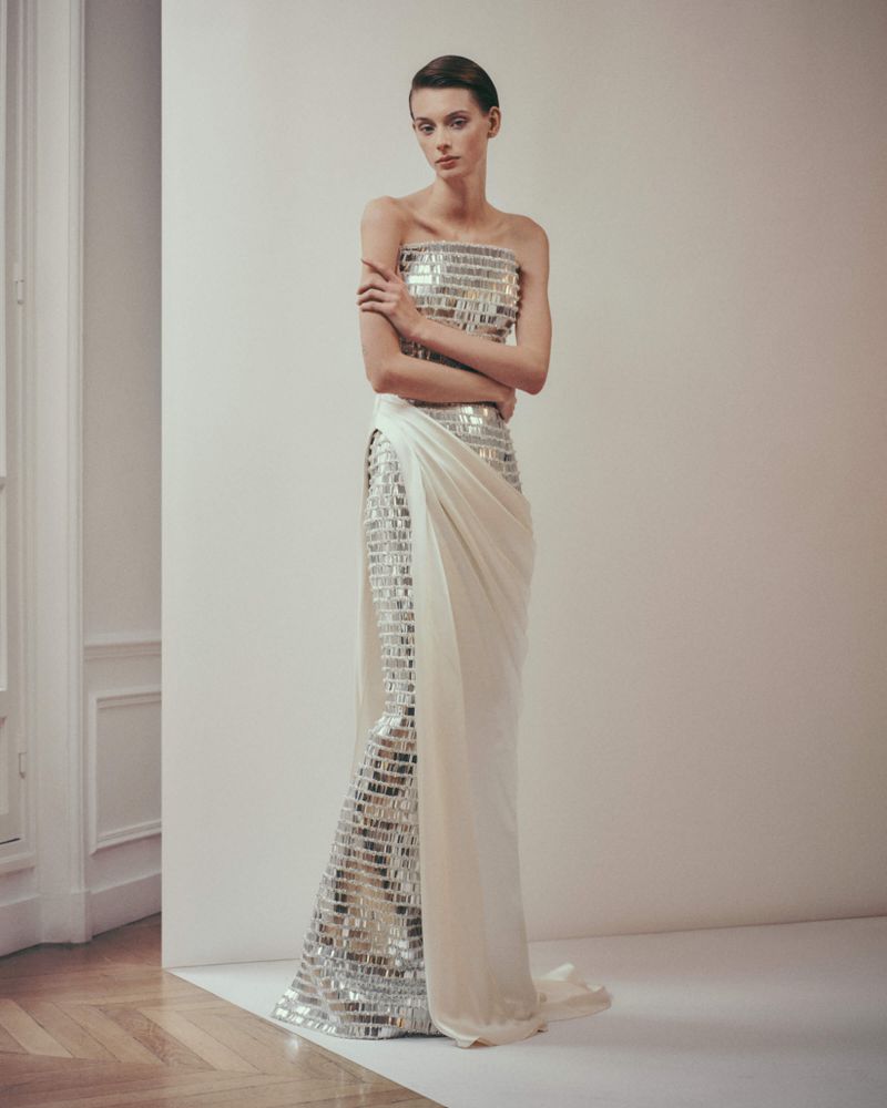 Elegant woman in a strapless, shimmering gown posing in an indoor setting with neutral colors and soft lighting, showcasing fashion.