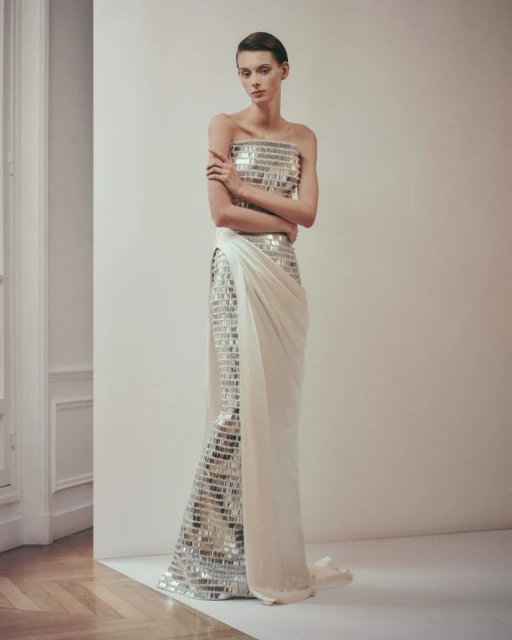 Elegant woman in a strapless, shimmering gown posing in an indoor setting with neutral colors and soft lighting, showcasing fashion.