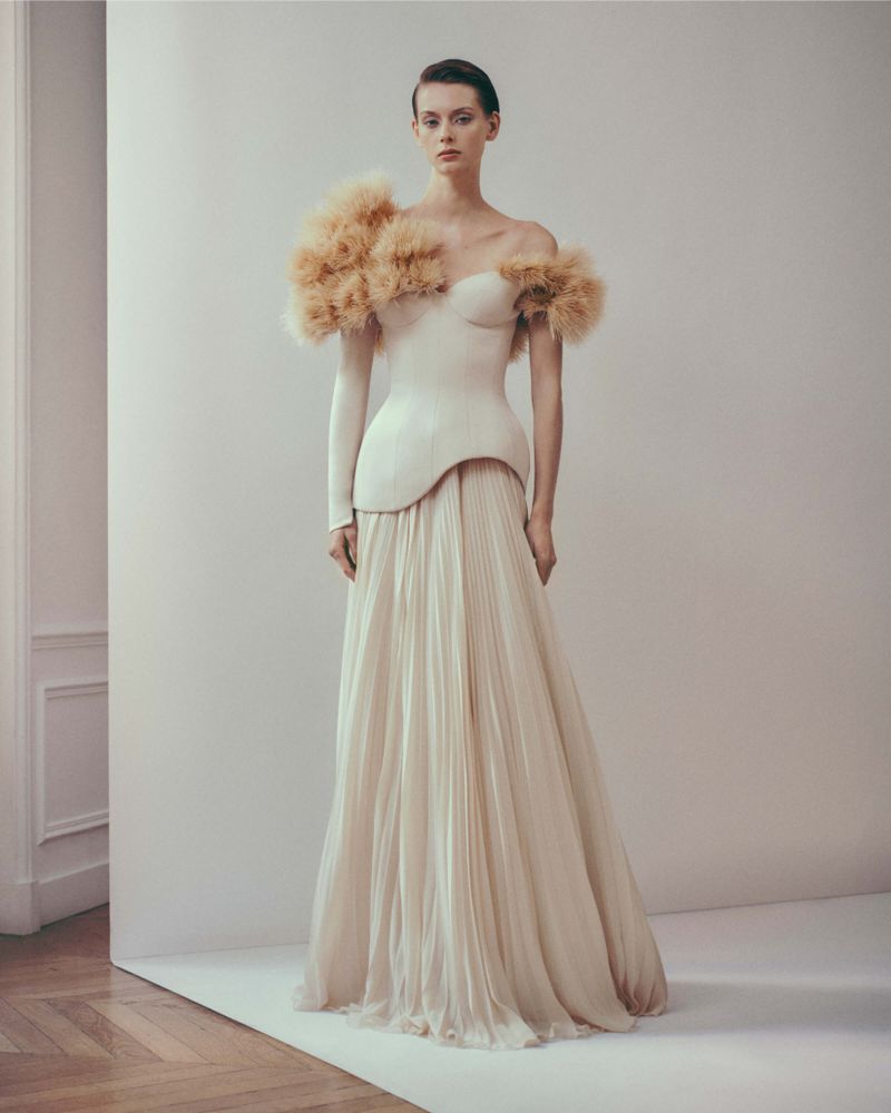 A model presents a white gown featuring textured shoulder embellishments, floor-length pleated skirt, and dramatic sleeve on a neutral set.