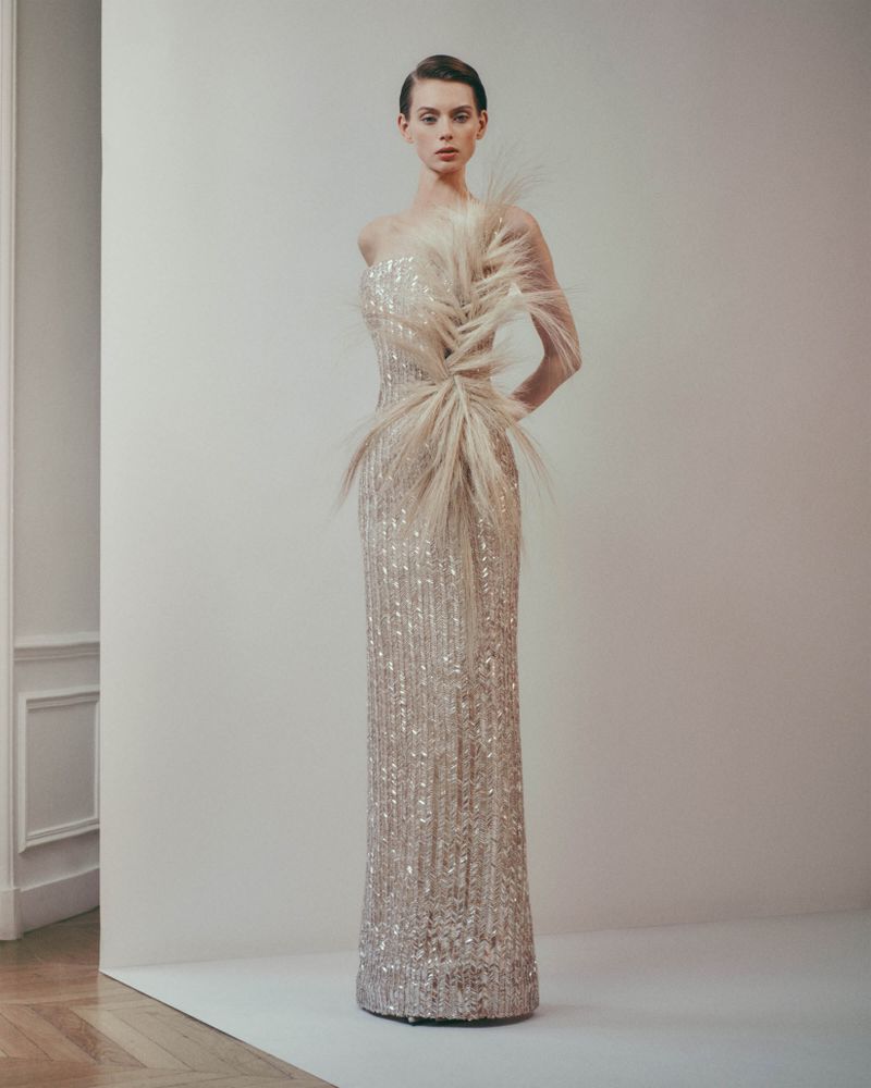 Elegant woman poses in sequined gown with feather embellishments in a neutral studio setting, fashion editorial, sophisticated style.