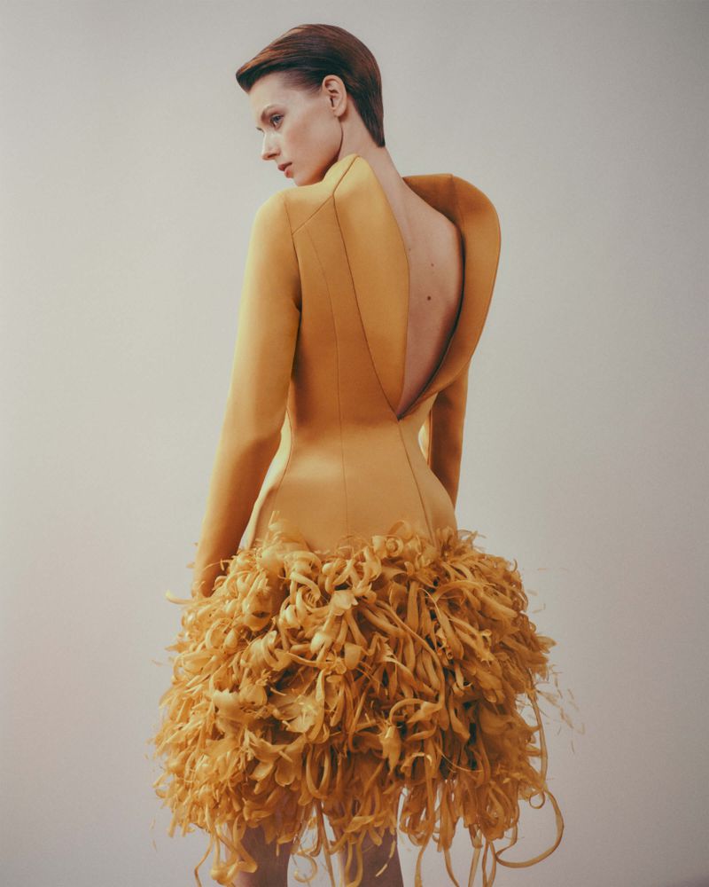 A slender adult female with short hair, wearing a gold dress with a feathered skirt, poses in a photo studio with soft lighting.