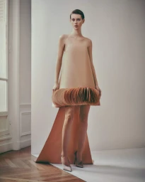 Model Lauren de Graaf in a sculptural nude strapless dress with an accordion pleated hem and train standing on a white backdrop.