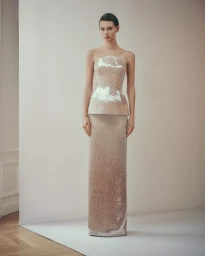 Model Lauren de Graaf wearing a strapless champagne sequin peplum evening gown against a pale studio backdrop for Ashi Studio.