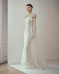 Model Lauren de Graaf in a white strapless beaded bridal gown with a sheer cape overlay against a minimalist neutral backdrop.