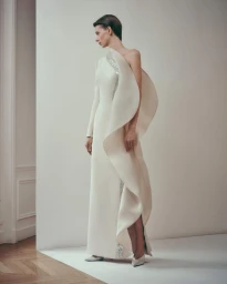 Model Lauren de Graaf standing in a sculptural off-white one-shoulder evening gown with curved ruffle detail against a white studio wall.