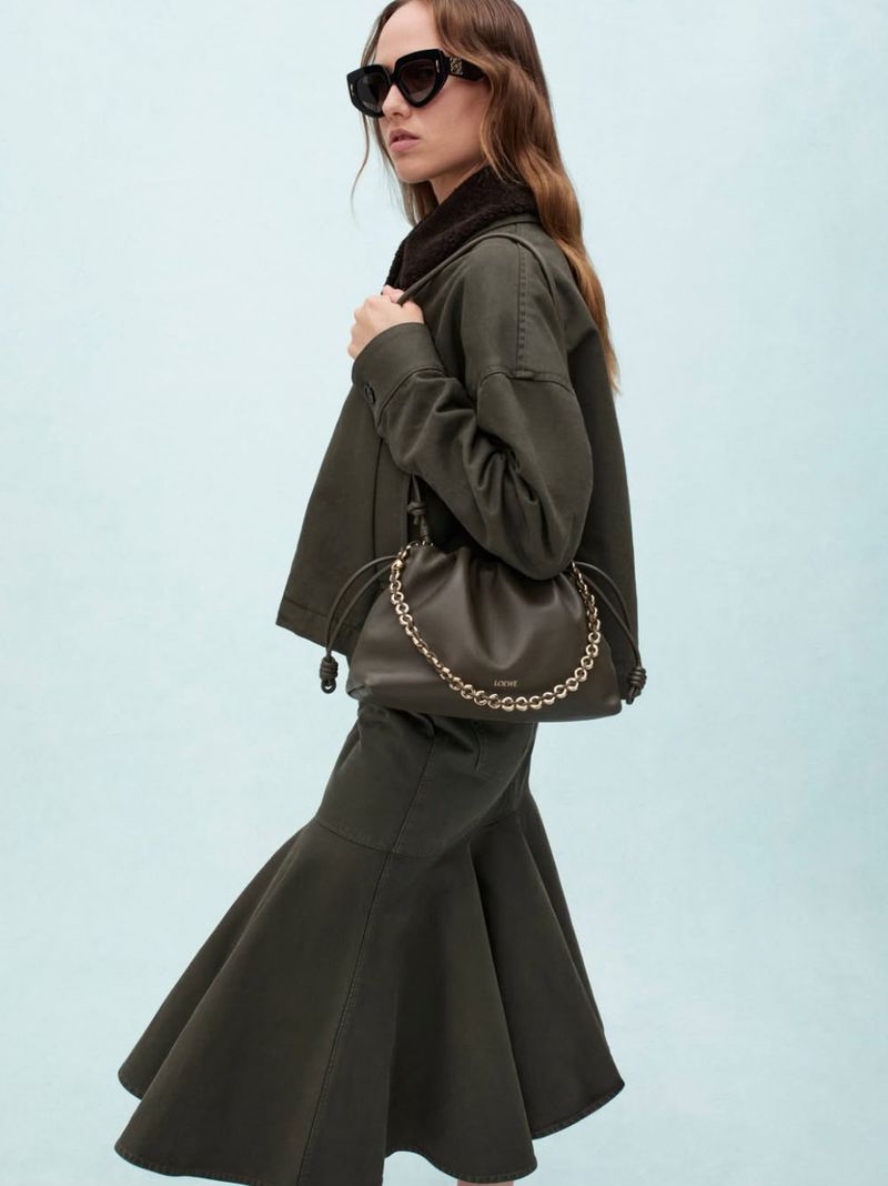 Model Hanna Felding wearing black sunglasses, an olive green jacket and flared skirt, holding an olive bag against a pale blue backdrop.