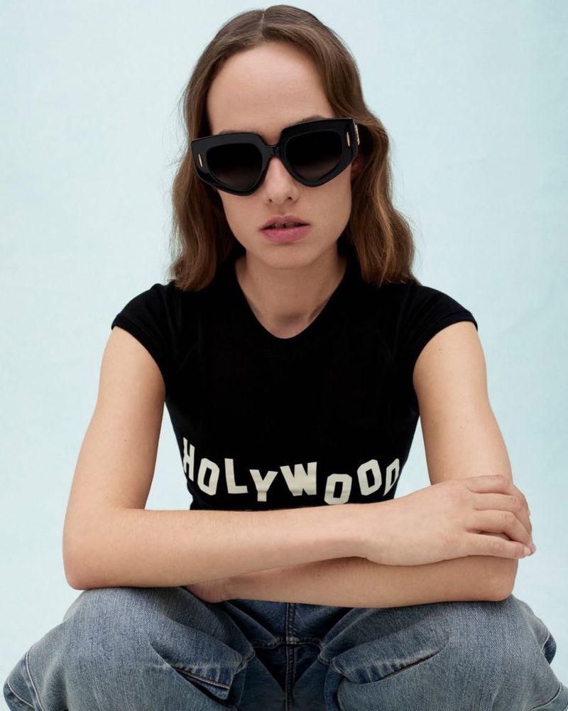 Model Hanna Felding wearing oversized black geometric sunglasses, a black graphic tee, and denim jeans against a light blue backdrop.