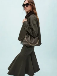 Model Hanna Felding wearing black sunglasses, an olive green jacket and flared skirt, holding an olive bag against a pale blue backdrop.