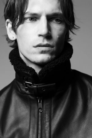 Close-up studio portrait of a fair-skinned adult male wearing a black leather jacket with a shearling collar, looking off camera.