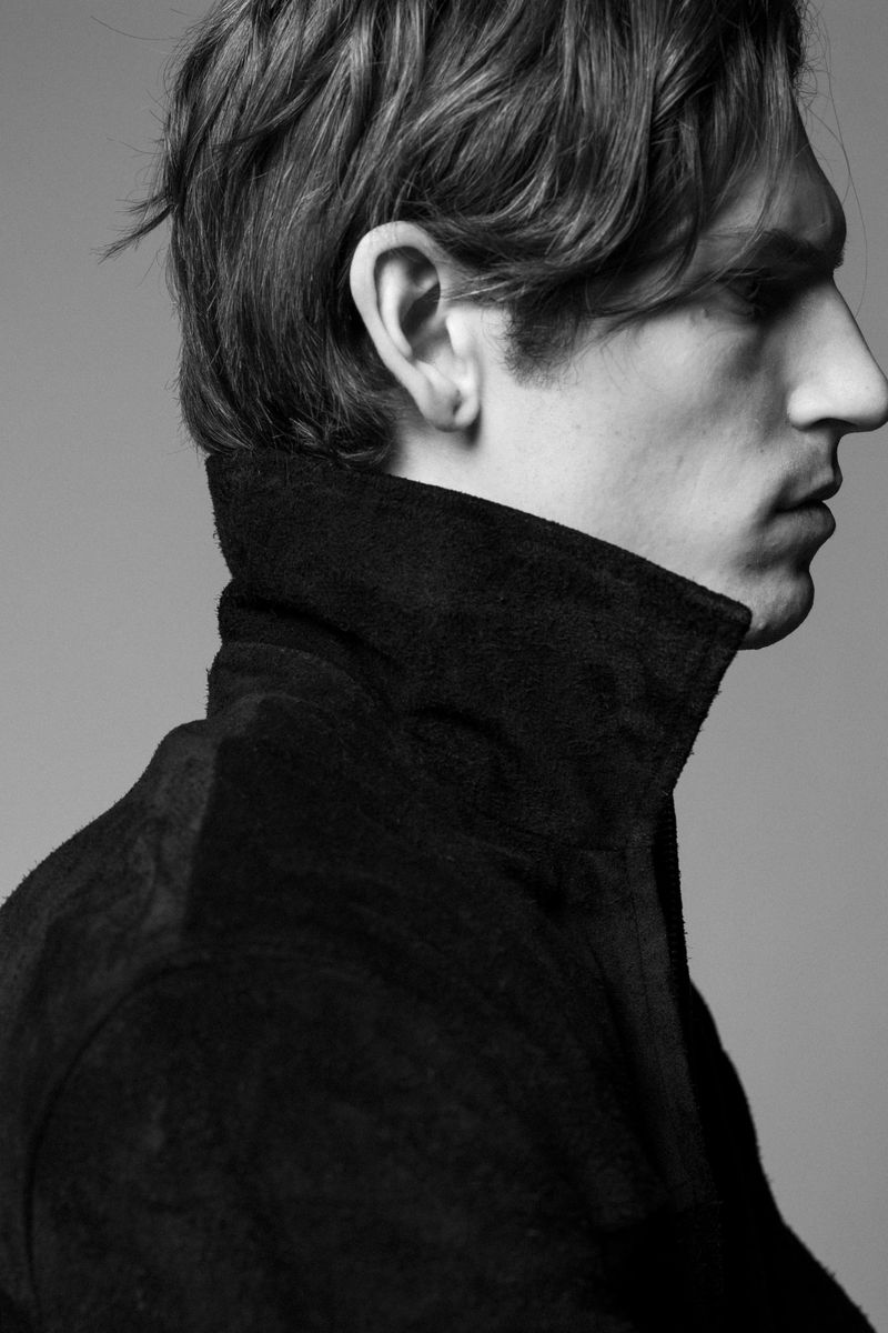 Monochrome portrait of a fair-skinned adult man in profile, facing right, wearing a black coat with a raised collar against a grey background.