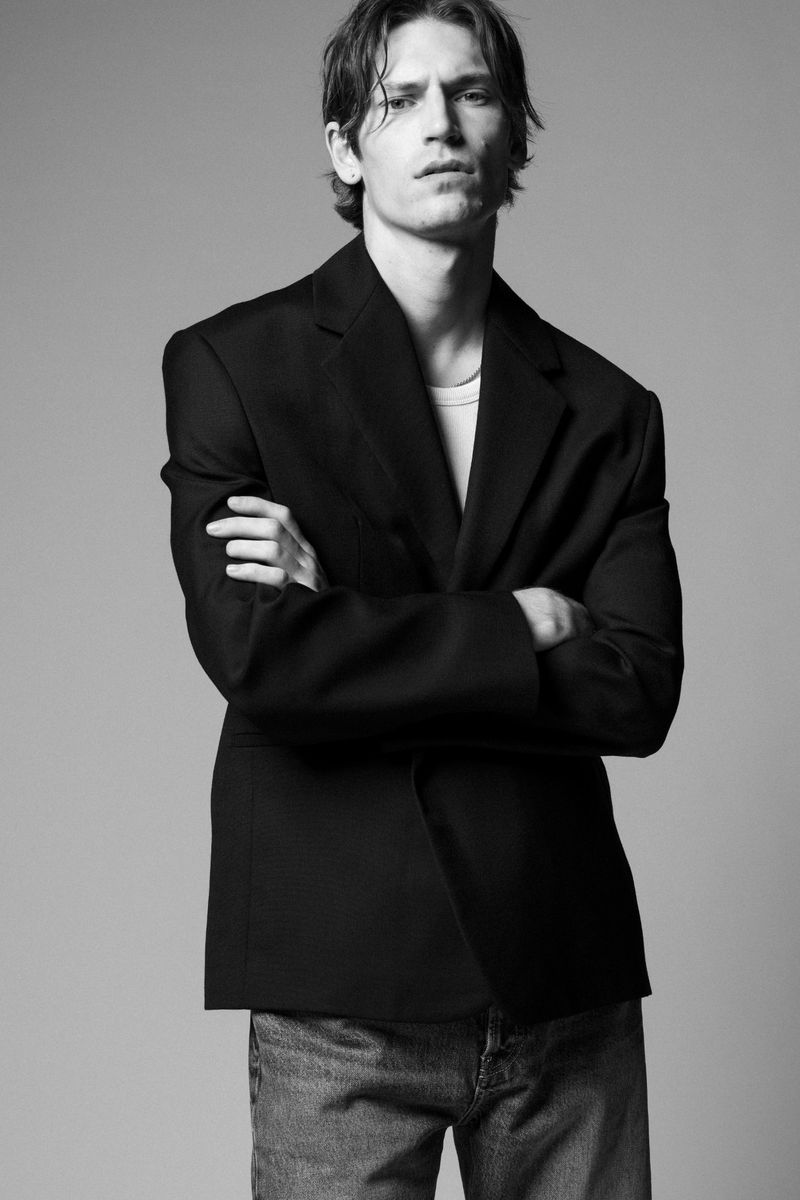Black-and-white portrait of a young adult male in a blazer and jeans with arms crossed against a neutral backdrop in a photo studio.