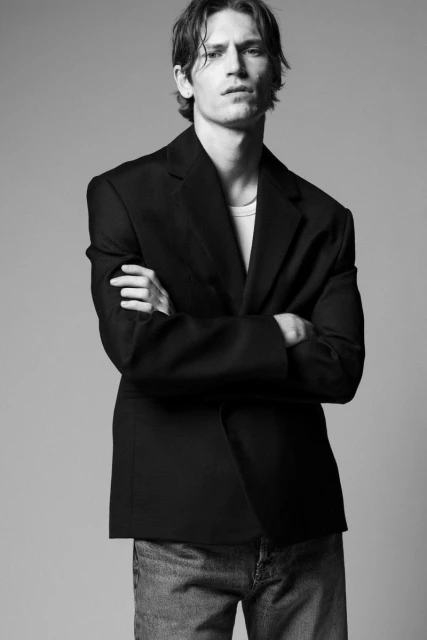 Black-and-white portrait of a young adult male in a blazer and jeans with arms crossed against a neutral backdrop in a photo studio.