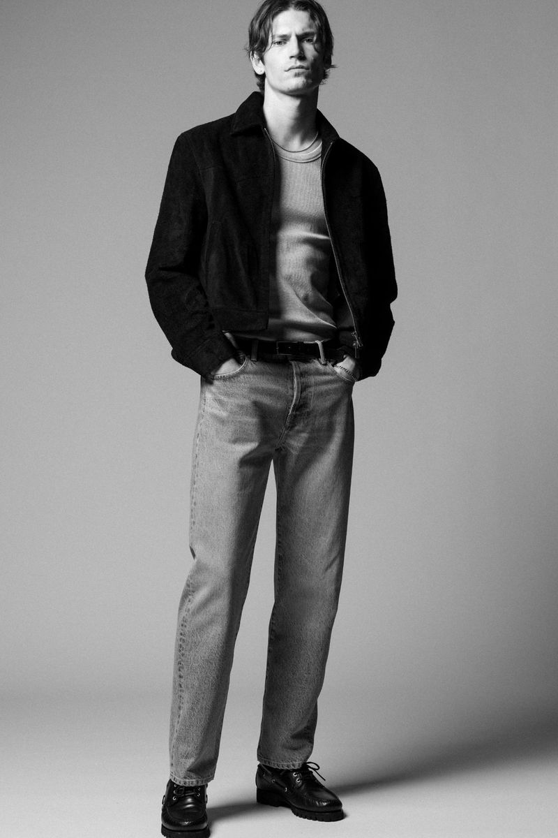 Full-length portrait of a male model in a suede jacket and jeans, posing indoors against a neutral gray backdrop in black and white.