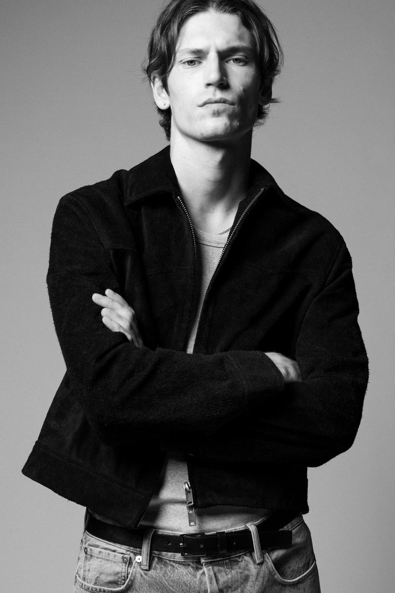 Black and white portrait of a young man wearing a jacket and jeans, standing with arms crossed in a photo studio setting.