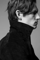 Black and white profile portrait of a man wearing a dark high-collar jacket against a plain studio background.
