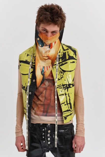 A person wears a graphic-print vest and a graphic-print face covering, standing in a bright studio against a white backdrop.