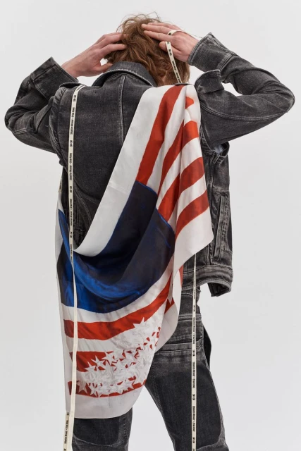 Back view of an androgynous person with red hair wearing a distressed denim jacket with an American flag draped over the shoulder.