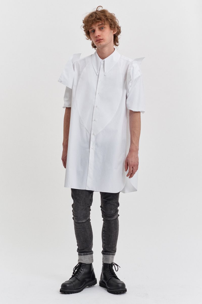 A full-length studio shot of a male model showcasing a long white shirt with angular details, paired with grey jeans and black boots.