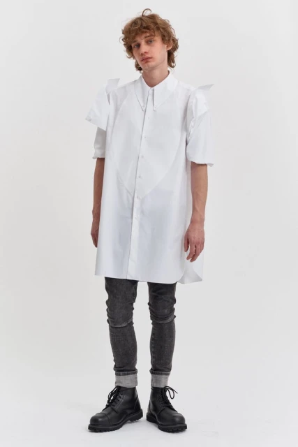 A full-length studio shot of a male model showcasing a long white shirt with angular details, paired with grey jeans and black boots.