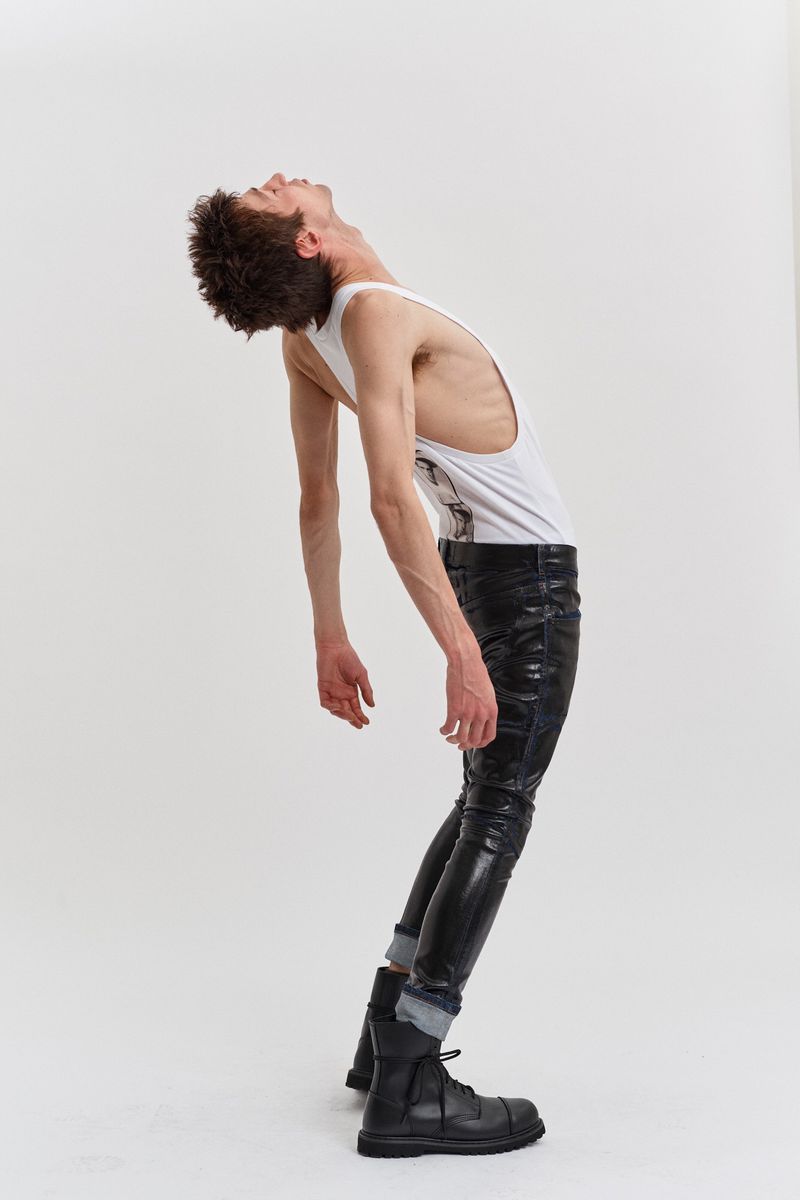 A slender man with curly hair in a white tank top, black pants, and boots arches his back against a plain white background in a fashion pose.