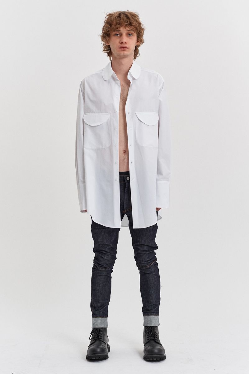 A full shot of a male model in a white oversized button-down shirt, dark jeans, and black boots against a plain white background.