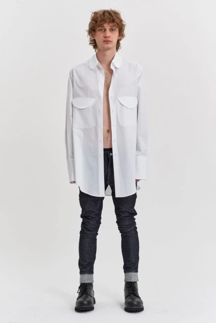 A full shot of a male model in a white oversized button-down shirt, dark jeans, and black boots against a plain white background.
