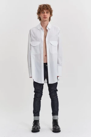 A full shot of a male model in a white oversized button-down shirt, dark jeans, and black boots against a plain white background.