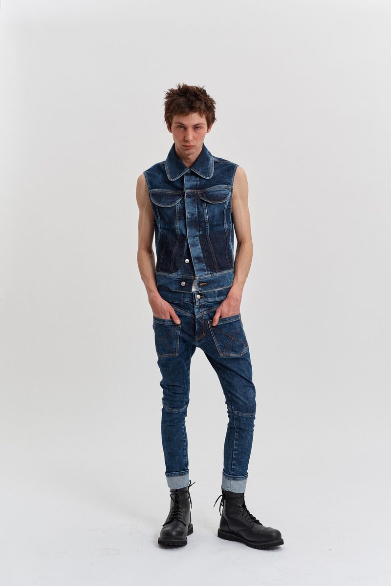 Full shot of a young adult man in denim overalls and black boots in a bright photo studio, posing with hands in pockets.
