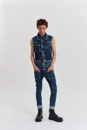 Full shot of a young adult man in denim overalls and black boots in a bright photo studio, posing with hands in pockets.