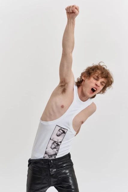 A young adult man with curly hair wearing a white tank top and black pants raises a fist in a photo studio against a white backdrop.