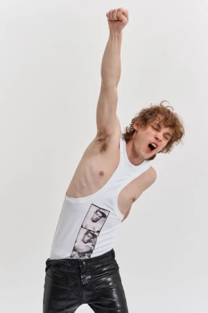 A young adult man with curly hair wearing a white tank top and black pants raises a fist in a photo studio against a white backdrop.