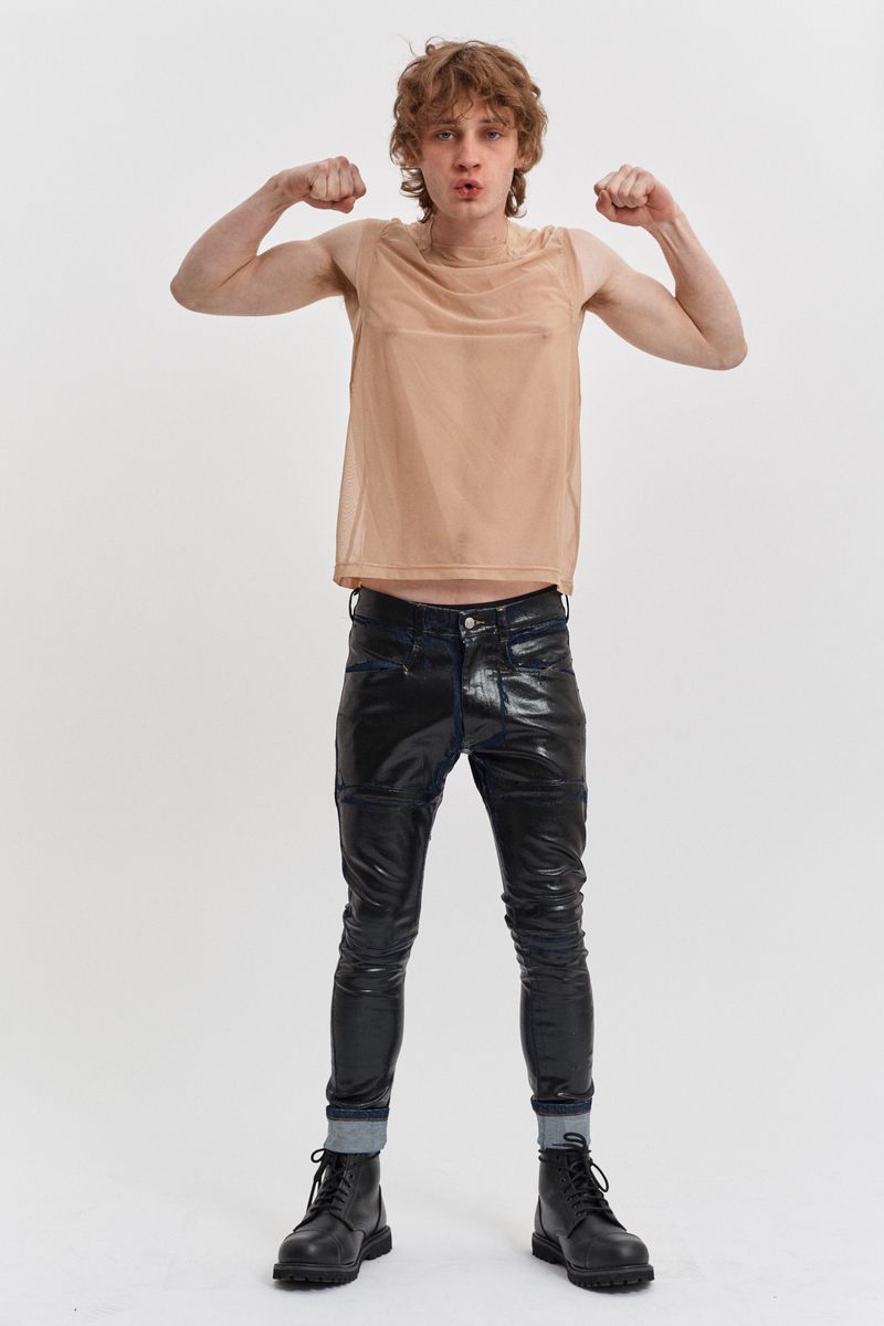 A male model poses in a studio, wearing a tan mesh tank top, black shiny pants, and black boots, flexing his arms with a neutral expression.
