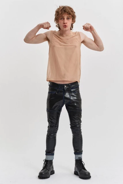 A male model poses in a studio, wearing a tan mesh tank top, black shiny pants, and black boots, flexing his arms with a neutral expression.