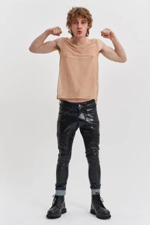 A male model poses in a studio, wearing a tan mesh tank top, black shiny pants, and black boots, flexing his arms with a neutral expression.