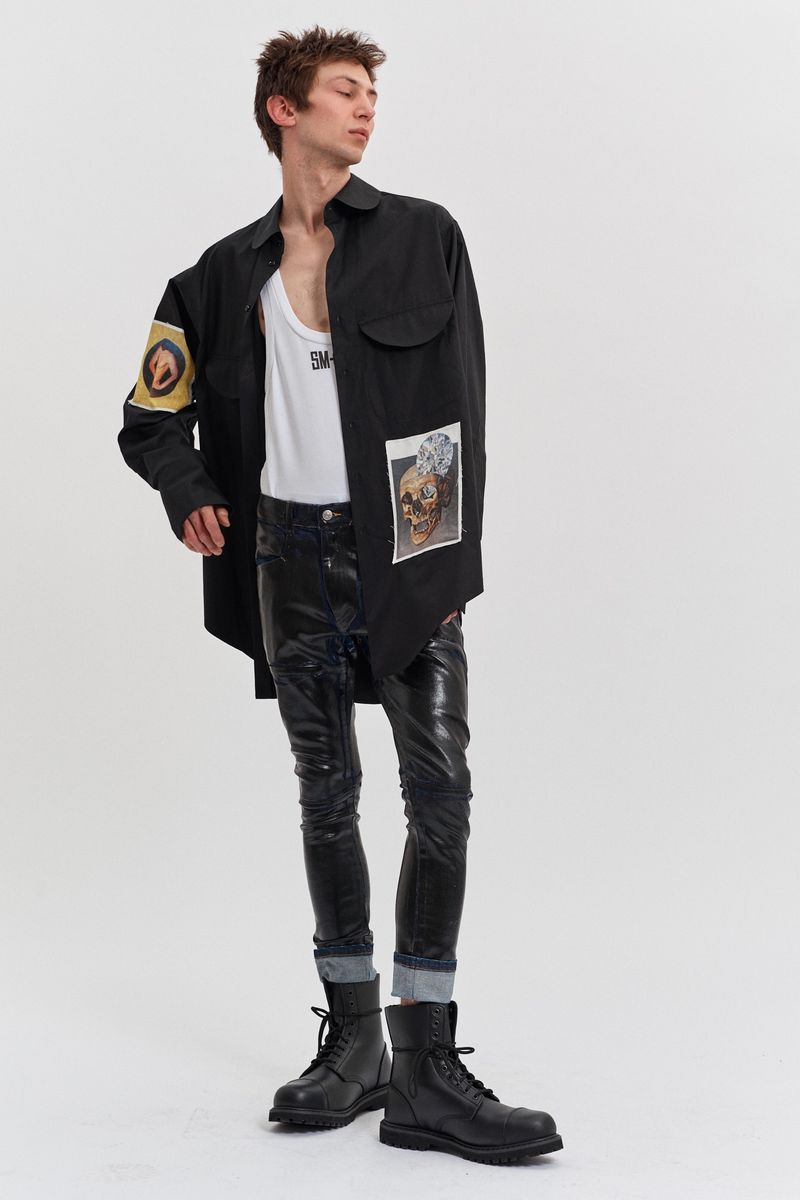 A slender male fashion model with tousled hair poses in a black shirt jacket, white tank top, dark jeans, and black combat boots against a white backdrop.