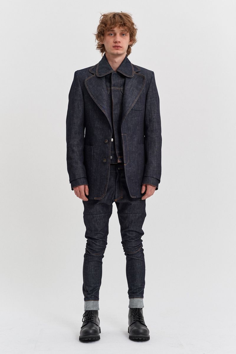 Full-length studio shot of a male fashion model in a dark-blue denim suit, shirt and jeans, posing against a white background.