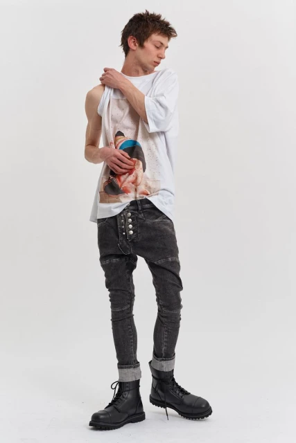 Full shot of a young adult man posing in a white tee shirt, acid-wash jeans with button details, and black boots on a white background.