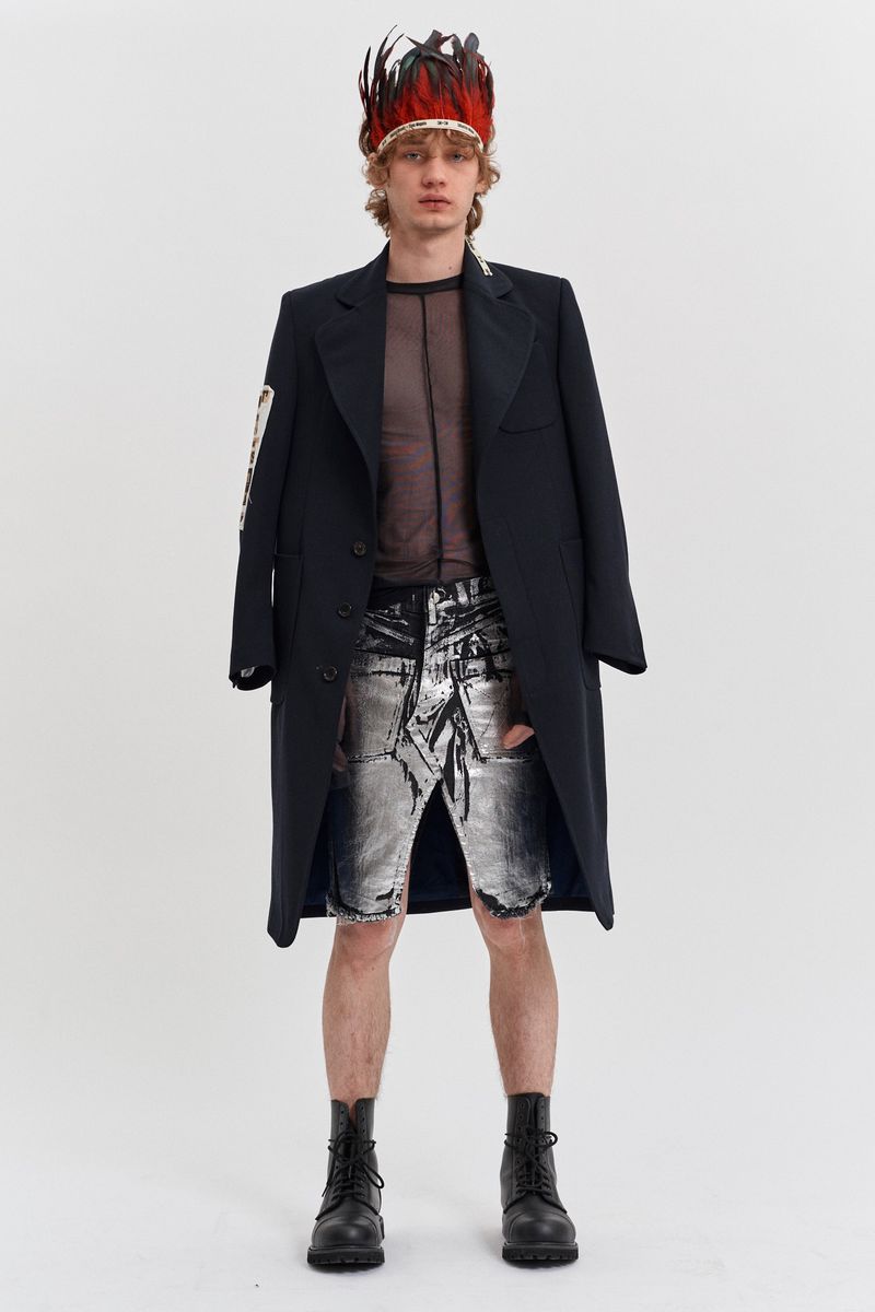 Male fashion model poses in a black coat, metallic shorts, black boots, and a feathered headdress against a plain white background.