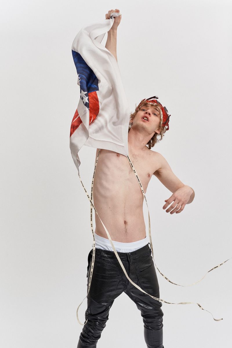 Young adult male with headdress stands shirtless in studio with American flag sweater in hand and LED lights around neck.