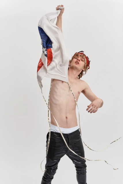 Young adult male with headdress stands shirtless in studio with American flag sweater in hand and LED lights around neck.